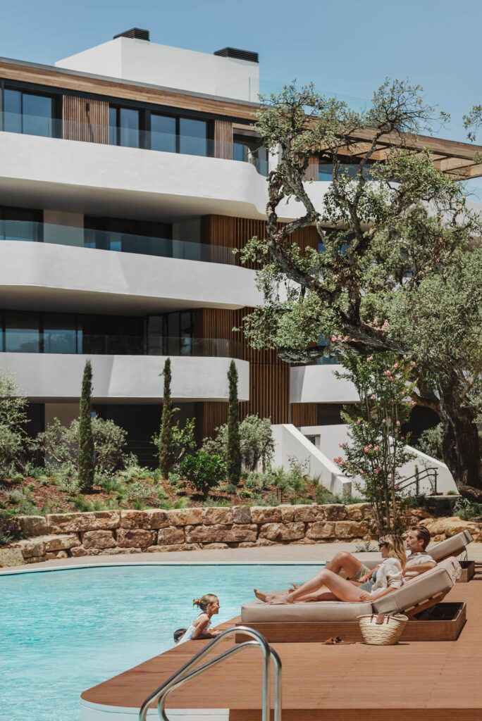 alt:"A photo of a family lying on a poolside hammock, with a building from Village Verde of Sotogrande in the background.Lifestyle Photography"